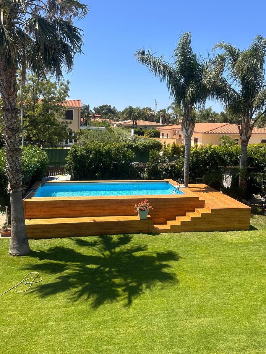 La piscina rivestita in legno vista dal giardino, tra le palme e il prato